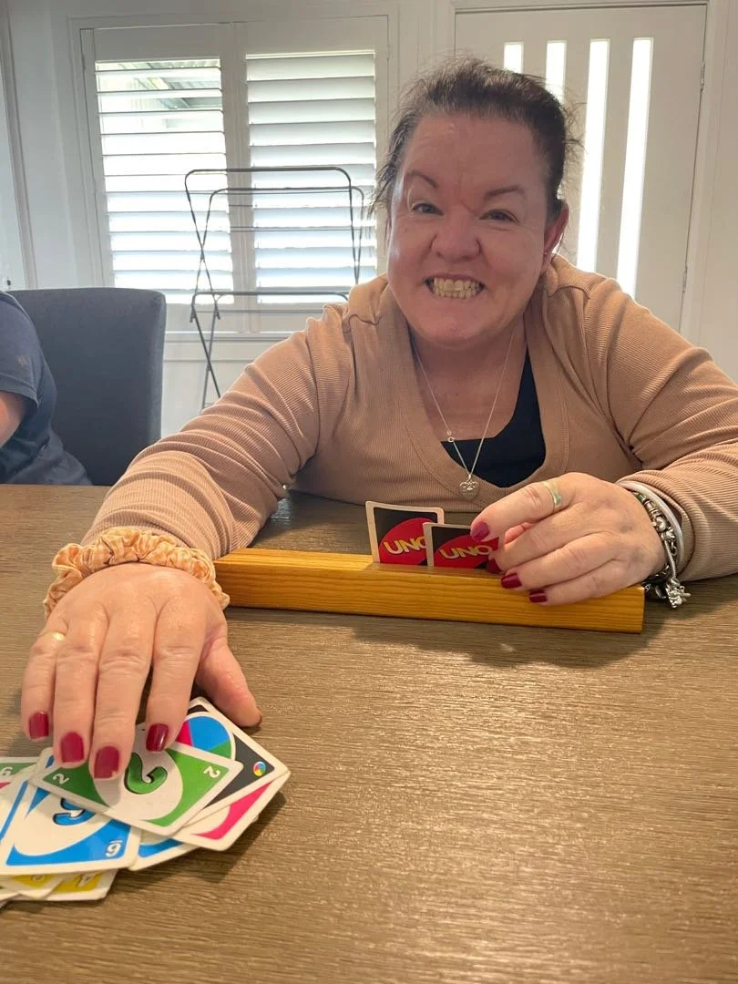 A woman playing UNO card game, holding cards in one hand, with an excited and playful facial expression, at a wooden table in a bright room with white shutters.