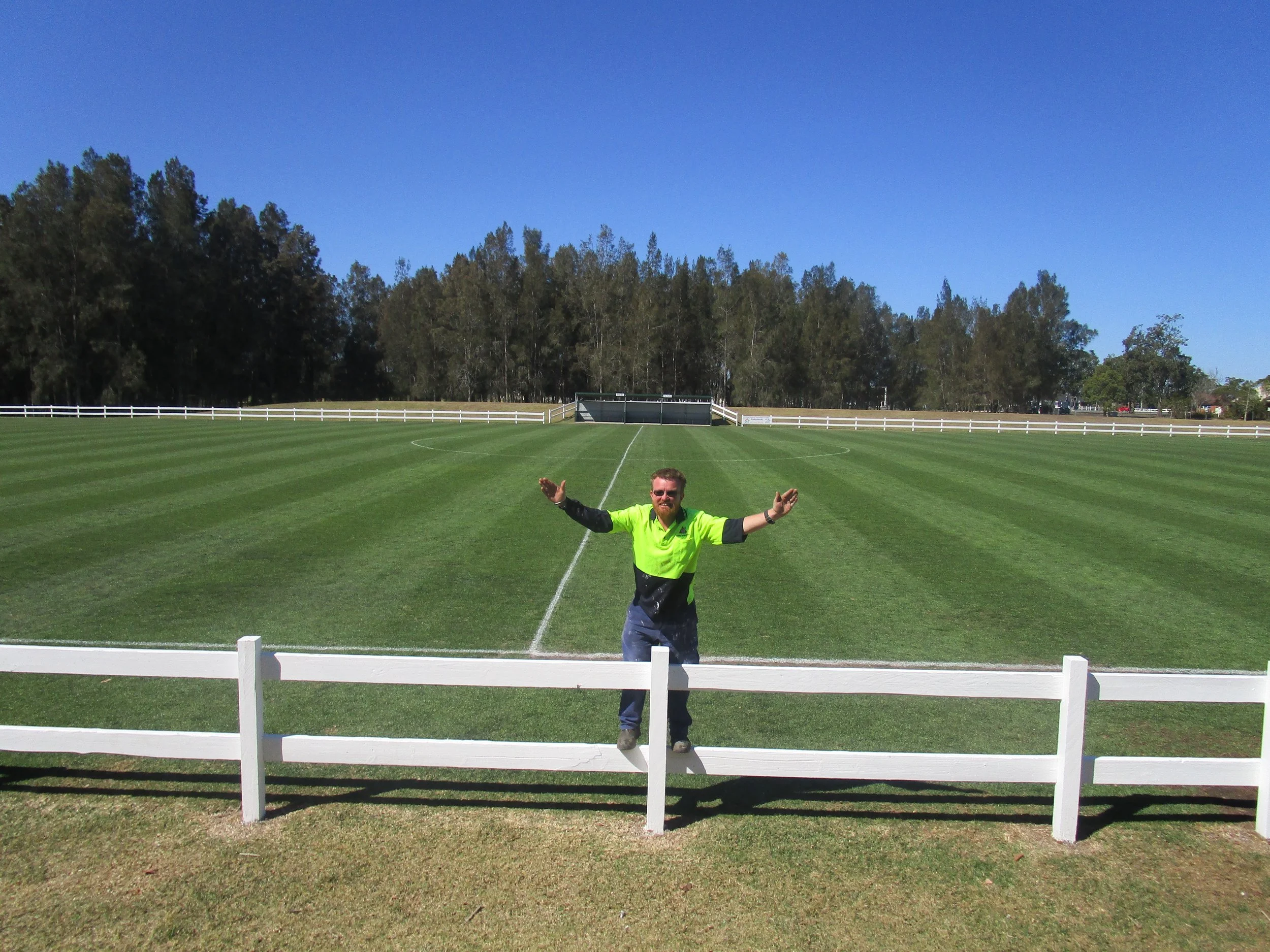 Man in a bright green shirt standing on a white fence on a football field with trees and blue sky in the background.