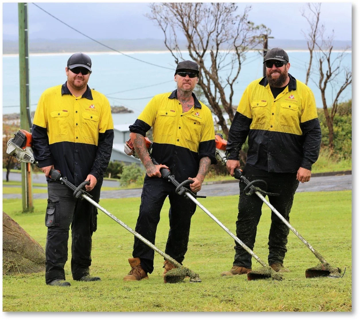Three workers in yellow and black uniforms using grass trimmers on a lawn near a coastal area with trees and water in the background.