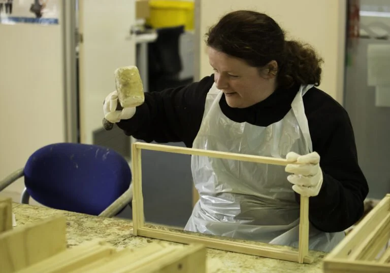 Woman wearing gloves and a plastic apron working on a woodworking project in a workshop, holding a piece of material and a wooden frame.