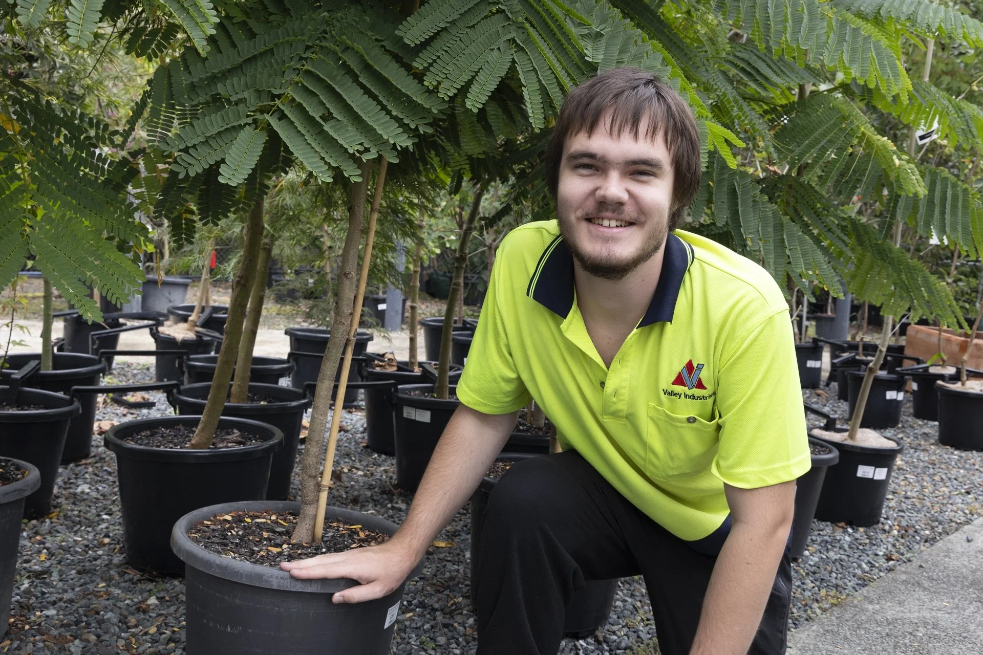 Young man in a bright‑yellow Valley Industries polo kneeling among potted trees at the Natures Care Nursery, learning safe woodworking techniques.