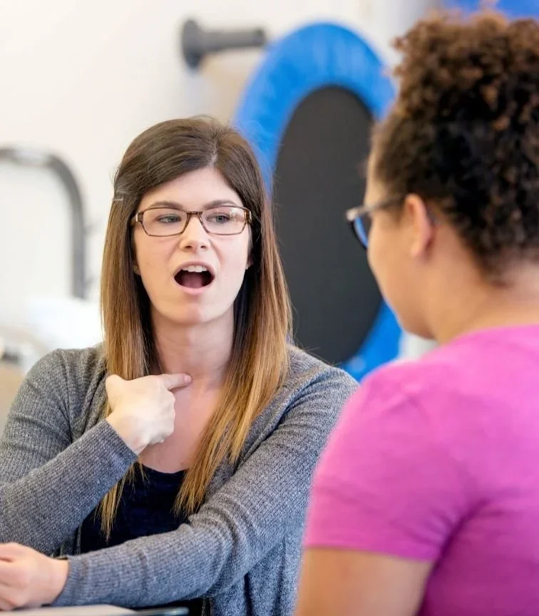 Two women engaged in an speech therapy session, one gesturing with her hand near her neck, wearing glasses and a gray sweater, the other wearing glasses and a pink shirt, in an indoor setting with a large blue exercise ball in the background.