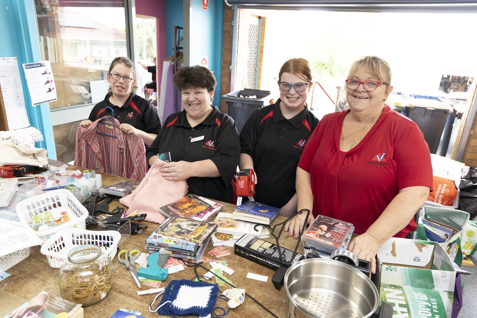 Four Valley Vintage team members at a table with books, DVDs, scissors, and various items, smiling and indoors with large windows, some wearing black shirts with a logo, and one woman in a red shirt.