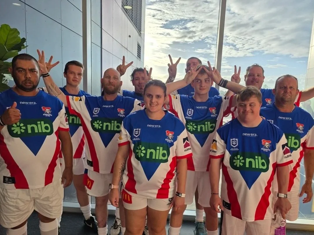 A group of rugby players in blue, red, and white jerseys making peace signs and posing for a team photo indoors with large windows showing a blue sky with clouds outside.