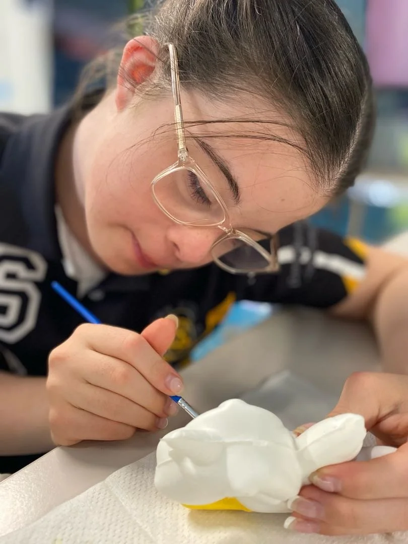 A young girl with glasses and brown hair working on a white ceramic figurine, painting it with a small brush.