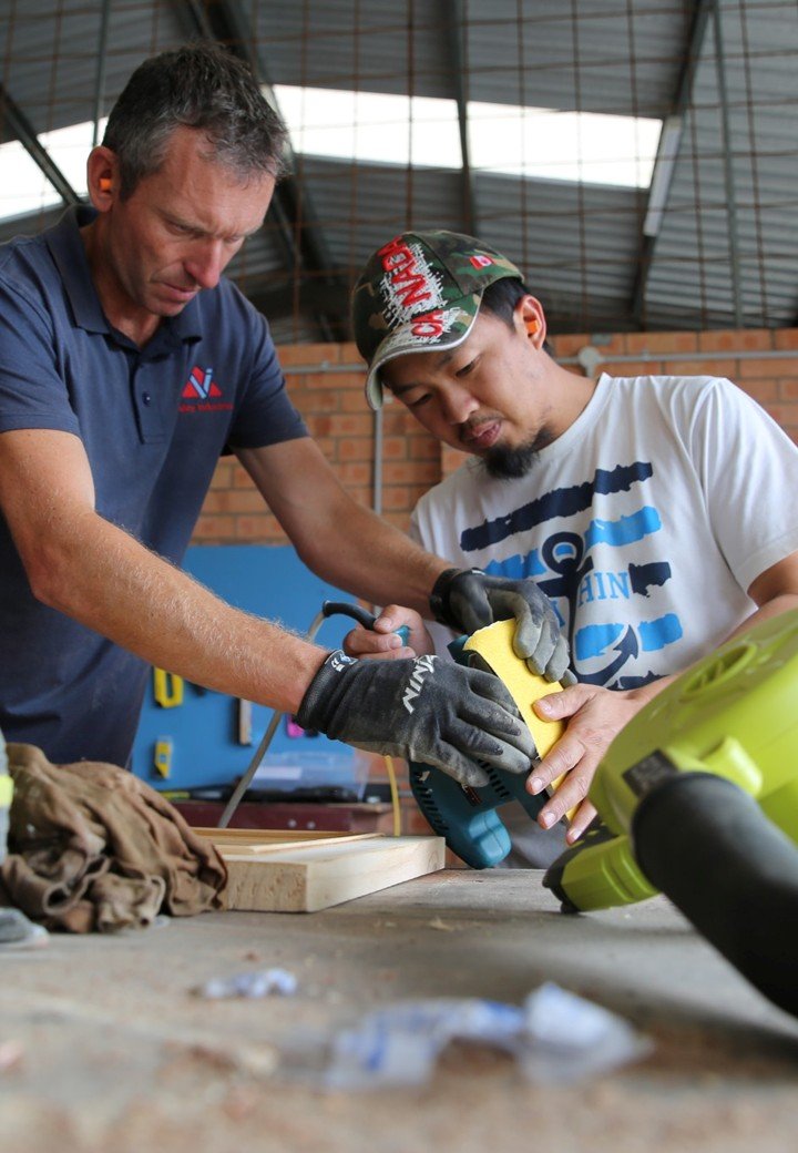 Two men working together on a woodworking project in a workshop. One man is guiding the other as they cut a piece of wood.