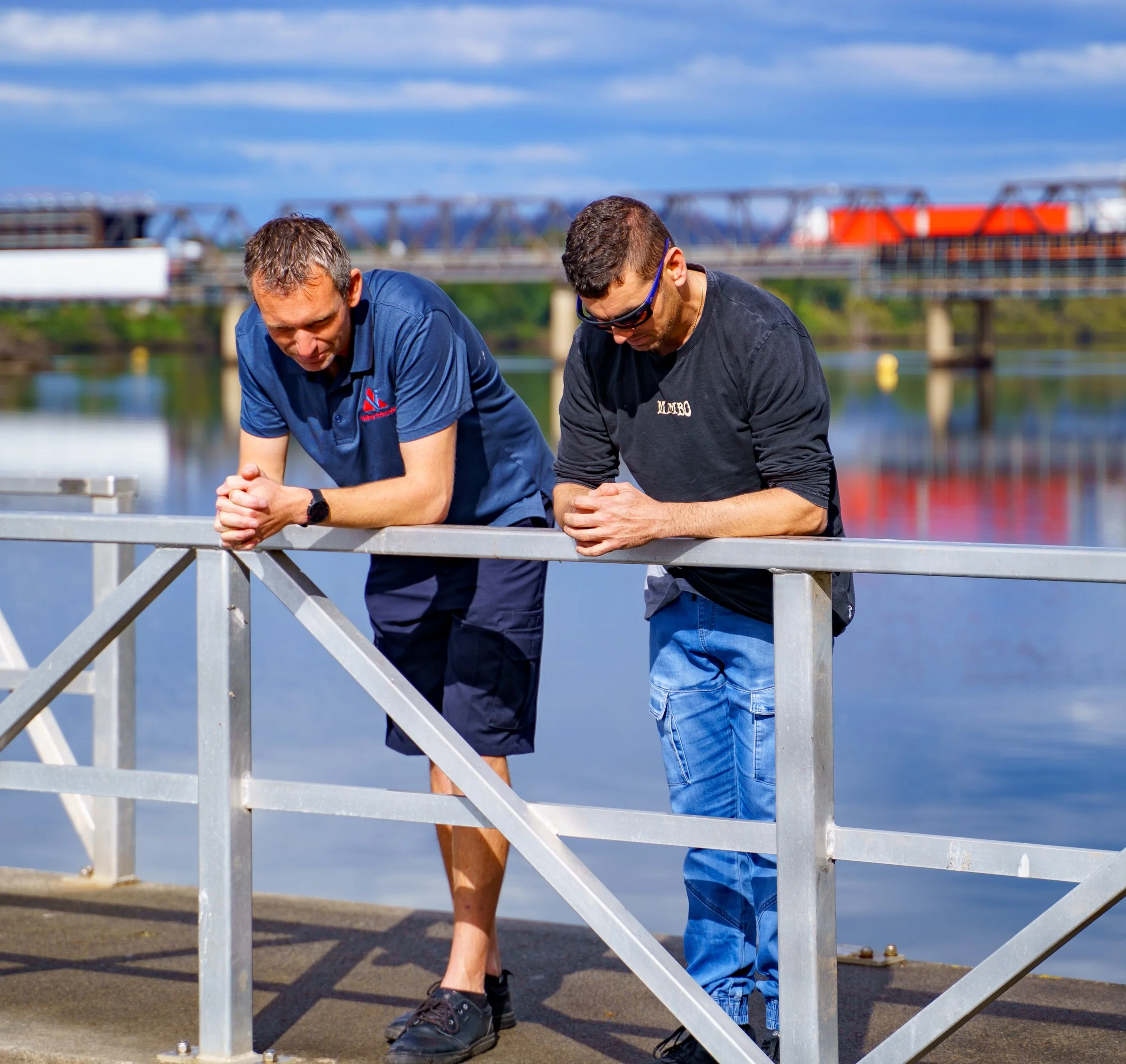 Two men leaning on a metal railing by a body of water during daytime, with a bridge in the background.