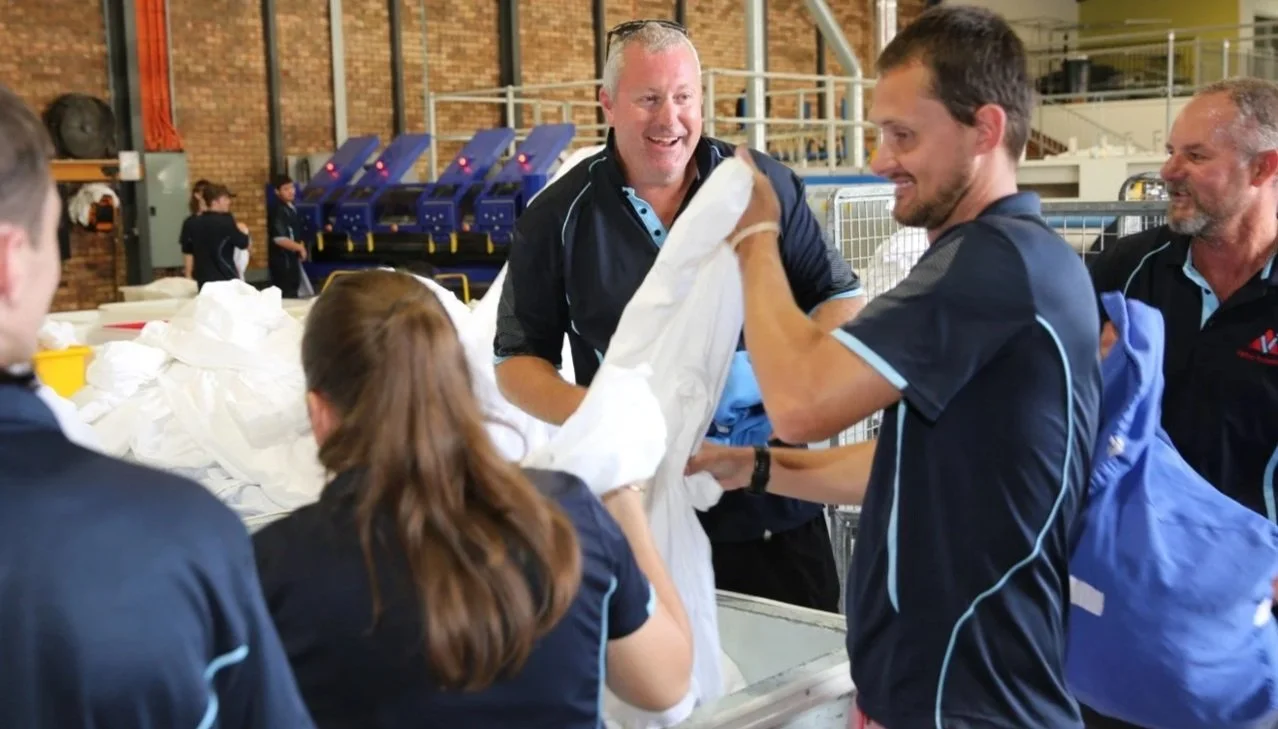 Group of people working together in an industrial setting, handling a large white fabric sheet, with some smiling and engaging with each other.
