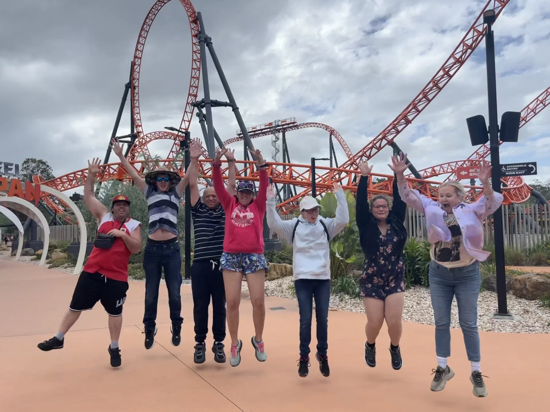 Group of seven people jumping in the air in front of a roller coaster at an amusement park.