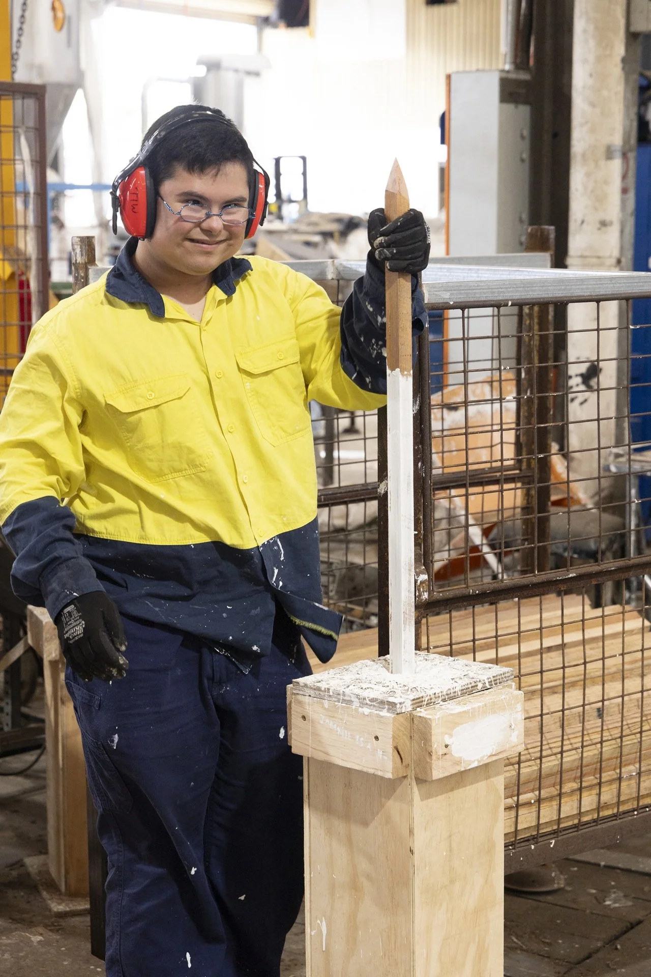 A young person wearing safety gear, including ear protection, glasses, and gloves, standing in a woodworking shop, holding a saw or chisel, with wood and metal fencing in the background.