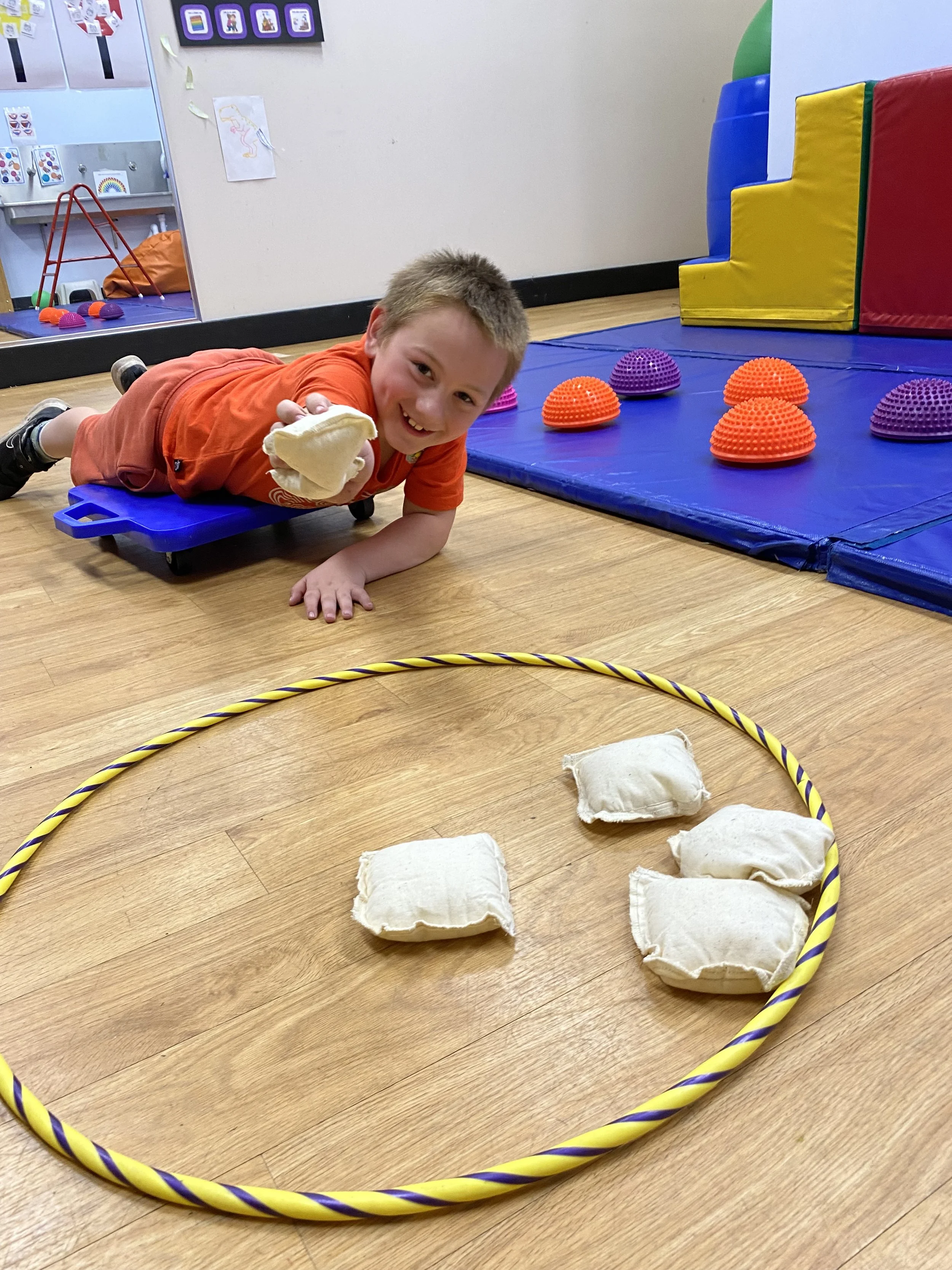 A young boy lying on a blue board on the floor of a playroom, smiling and holding a stuffed toy. In front of him on the wooden floor, there are four small fabric cushions inside a yellow and purple hula hoop. The background features colorful foam climbing blocks and other play equipment.