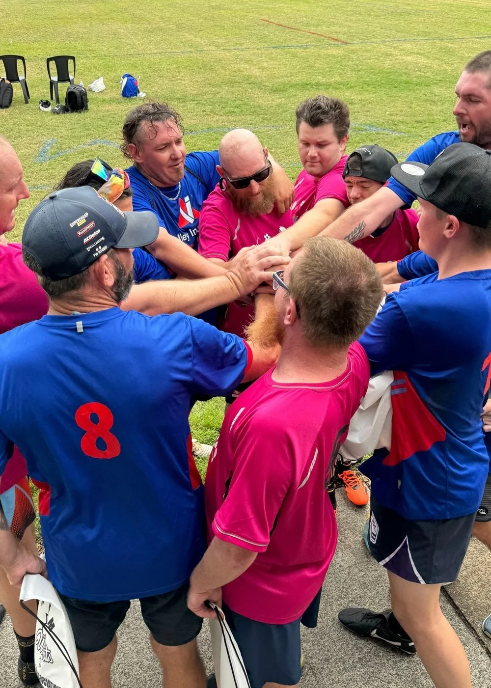 A group of athletes and coaches in pink and blue jerseys huddled together in a team cheer, hands stacked in the center, on a grassy sports field.