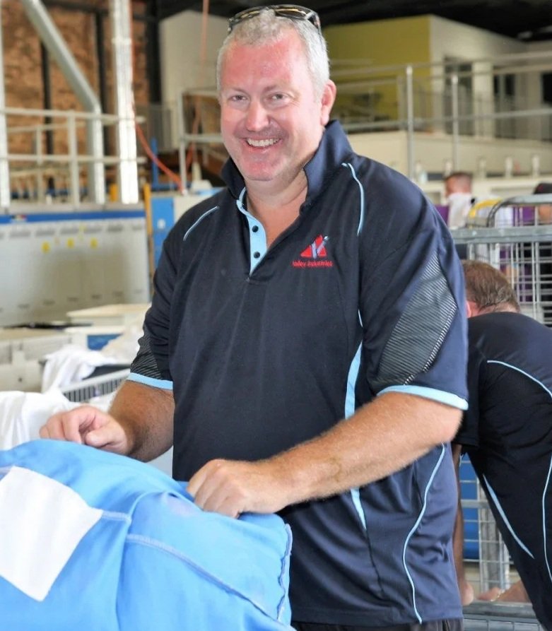 A smiling man with short gray hair and a beard, wearing a black athletic polo shirt with blue accents, standing in an industrial or warehouse setting.