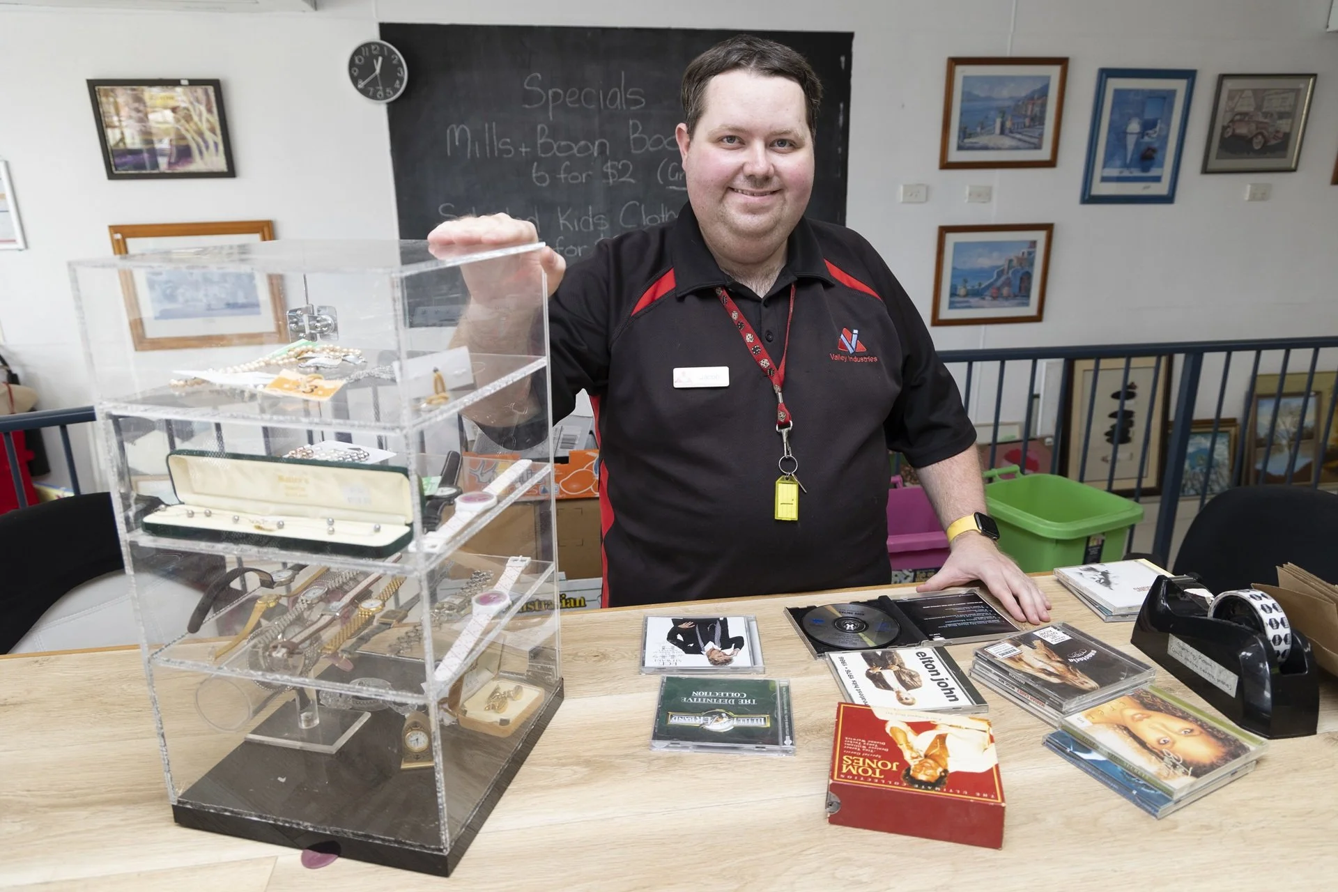 A man with short dark hair and a light complexion behind a table displays jewelry, CDs, and a tape dispenser, with framed pictures on the wall behind him and a chalkboard in the background in our Valley Vintage shop.