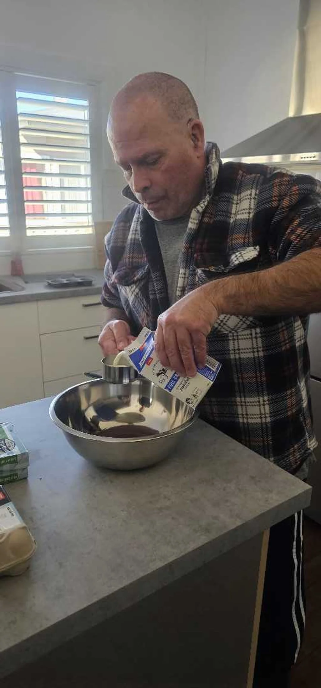 A man in a checkered shirt pouring milk into a mixing bowl in a kitchen.