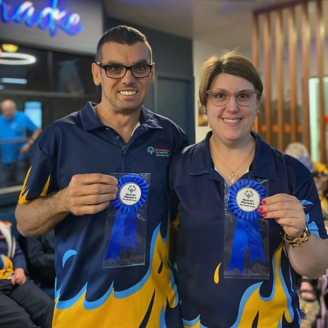 Two smiling individuals, a man and a woman, holding blue ribbon medals at a sporting event, wearing matching blue uniforms with yellow and blue flame designs.
