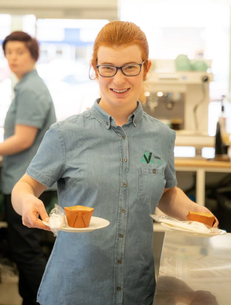 A smiling Valley team member with red hair and glasses, wearing a blue work shirt with a company logo, serving food on plates in our Valley TAFE Canteen.