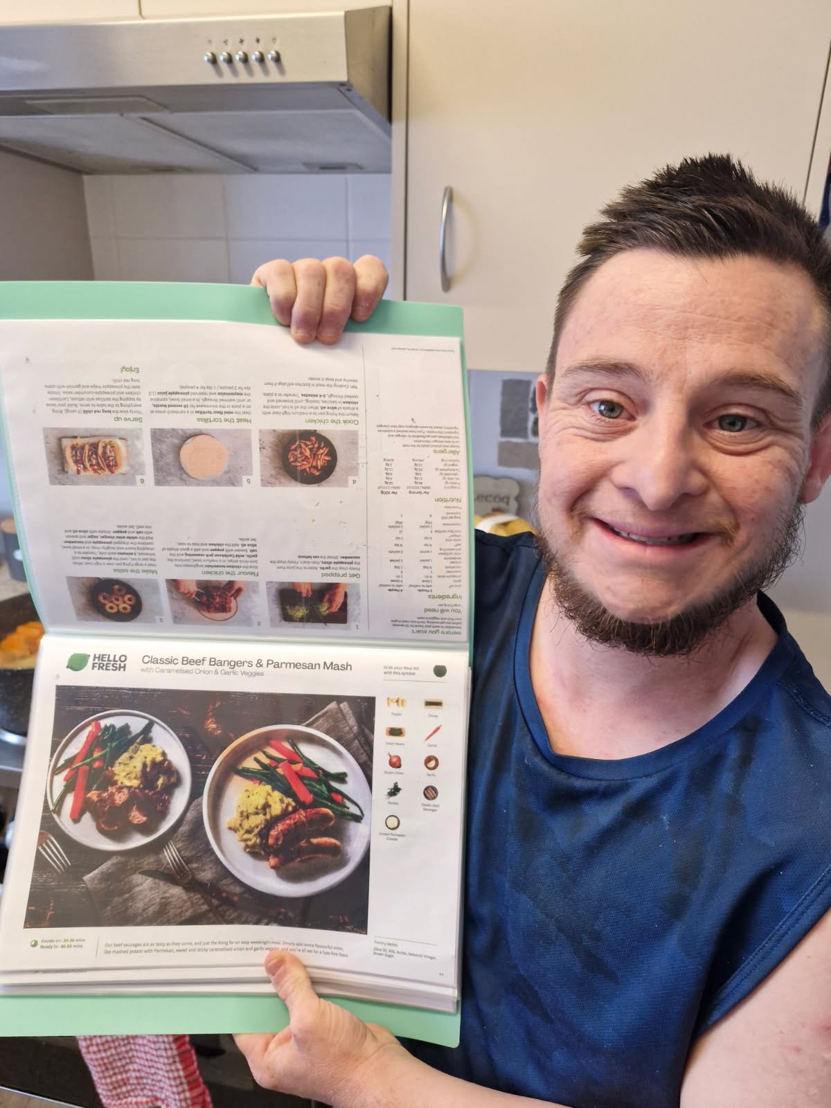 A smiling man holding a menu in front of a kitchen background.