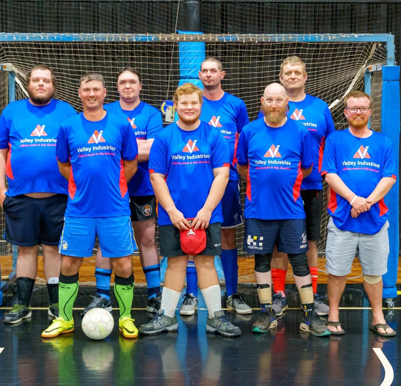 A group of nine people wearing blue sports jerseys with red accents, standing in front of a goal net on an indoor court, with a soccer ball on the floor in front of them.