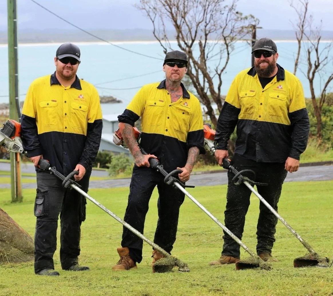 Three men in yellow and black work uniforms using leaf blowers on grass near a body of water and leafless trees.