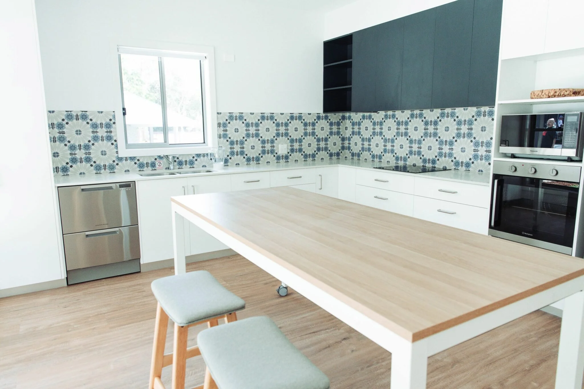Modern kitchen with white cabinets, patterned blue and white backsplash, stainless steel appliances, wooden dining table with light-colored stools, window with outside view.
