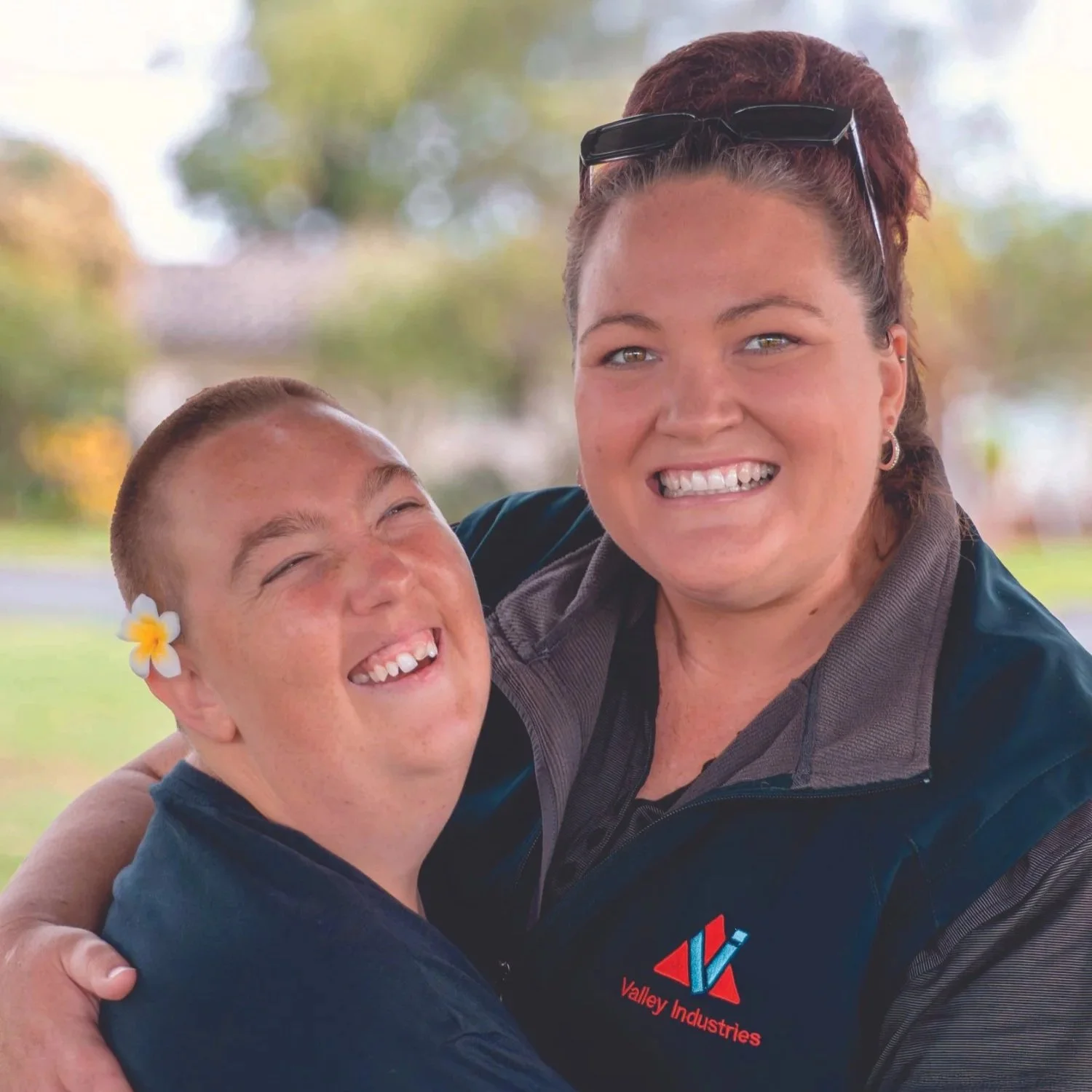 A Valley support worker hugging a client outdoors in Taree; both smile, the worker wears a Valley Industries jacket and the client has a flower behind the ear.