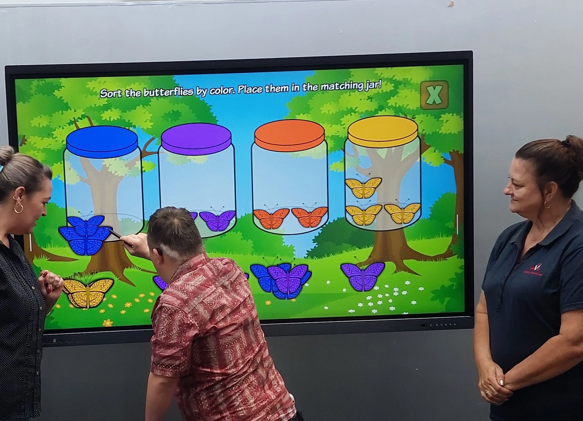 Children and adults look at a digital display with colorful butterflies and jars for sorting by color, in a park scene background.
