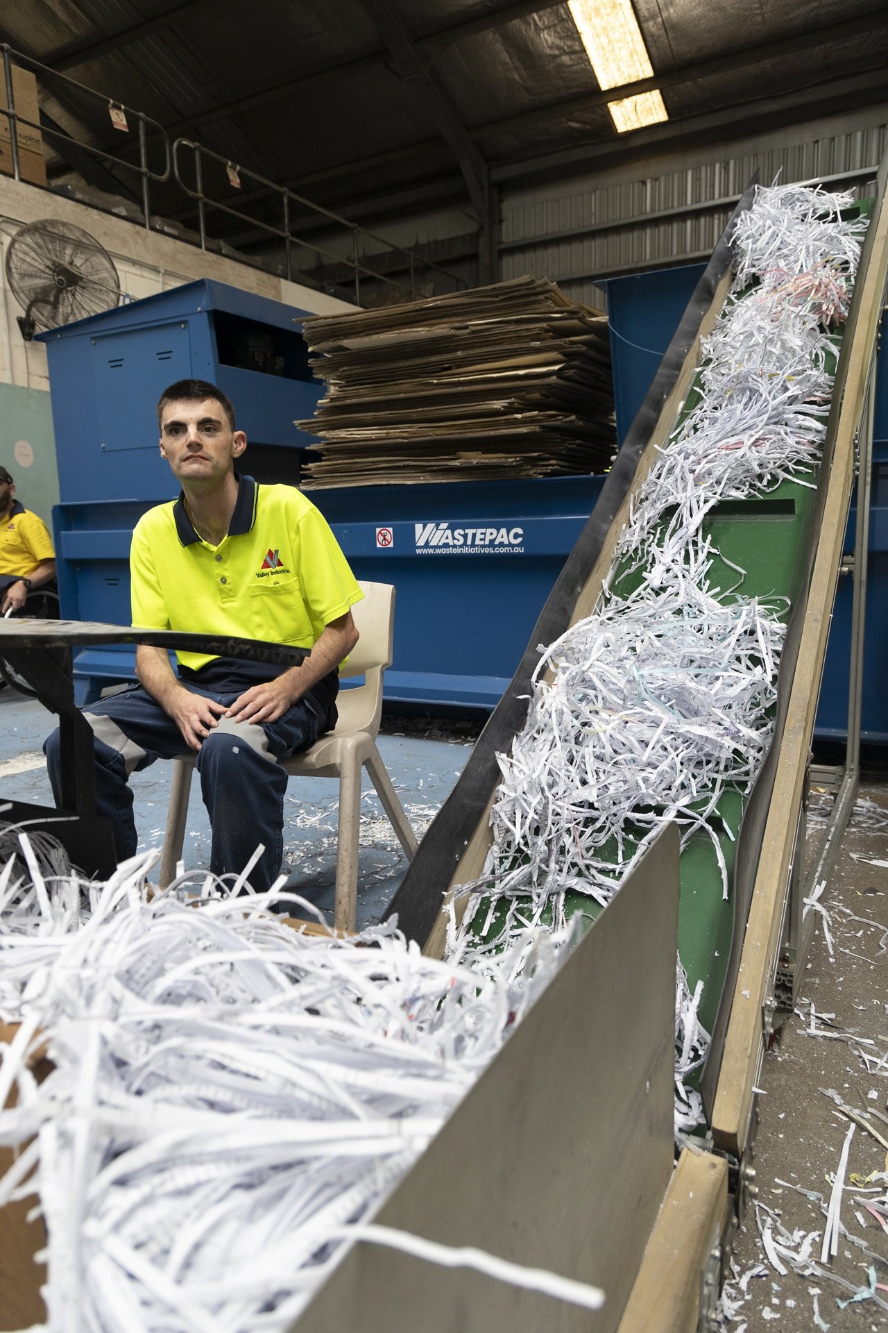 A man sits at a table in an industrial paper recycling facility, with shredded paper on a conveyor belt in the foreground and large blue recycling bins behind him.