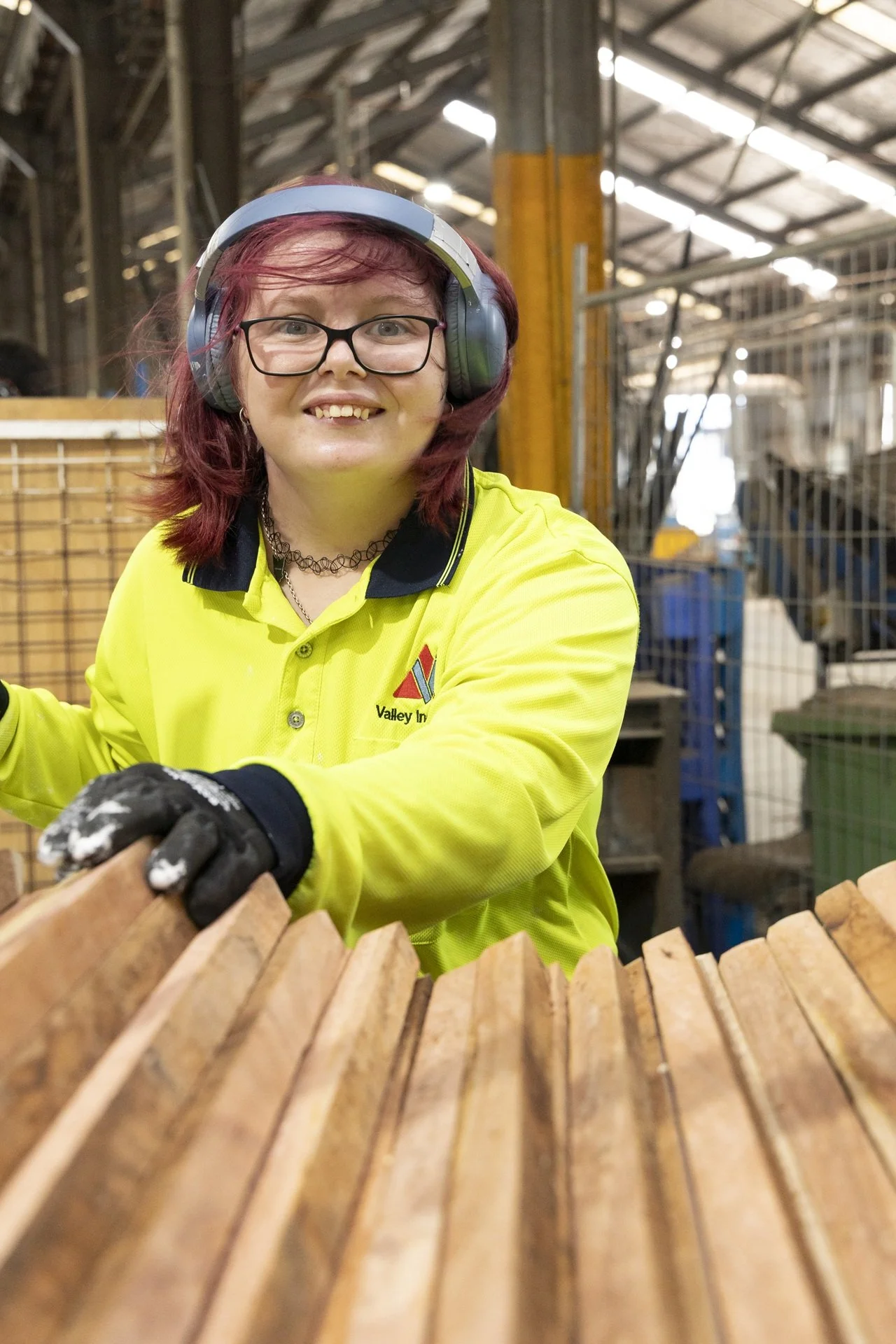 A woman wearing safety glasses, earmuffs, gloves, and a bright yellow high-visibility shirt smiling and working at a construction or woodworking site with wooden planks.