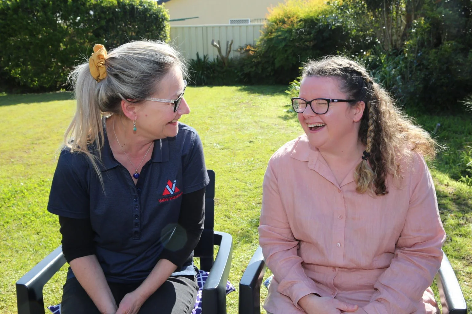 Two women sitting outside on chairs, sharing a laugh and smiling at each other in a garden with greenery and sunlight.