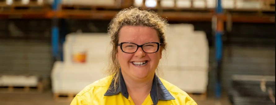A woman with curly hair, glasses, and a big smile, wearing a yellow work uniform, standing in a warehouse.