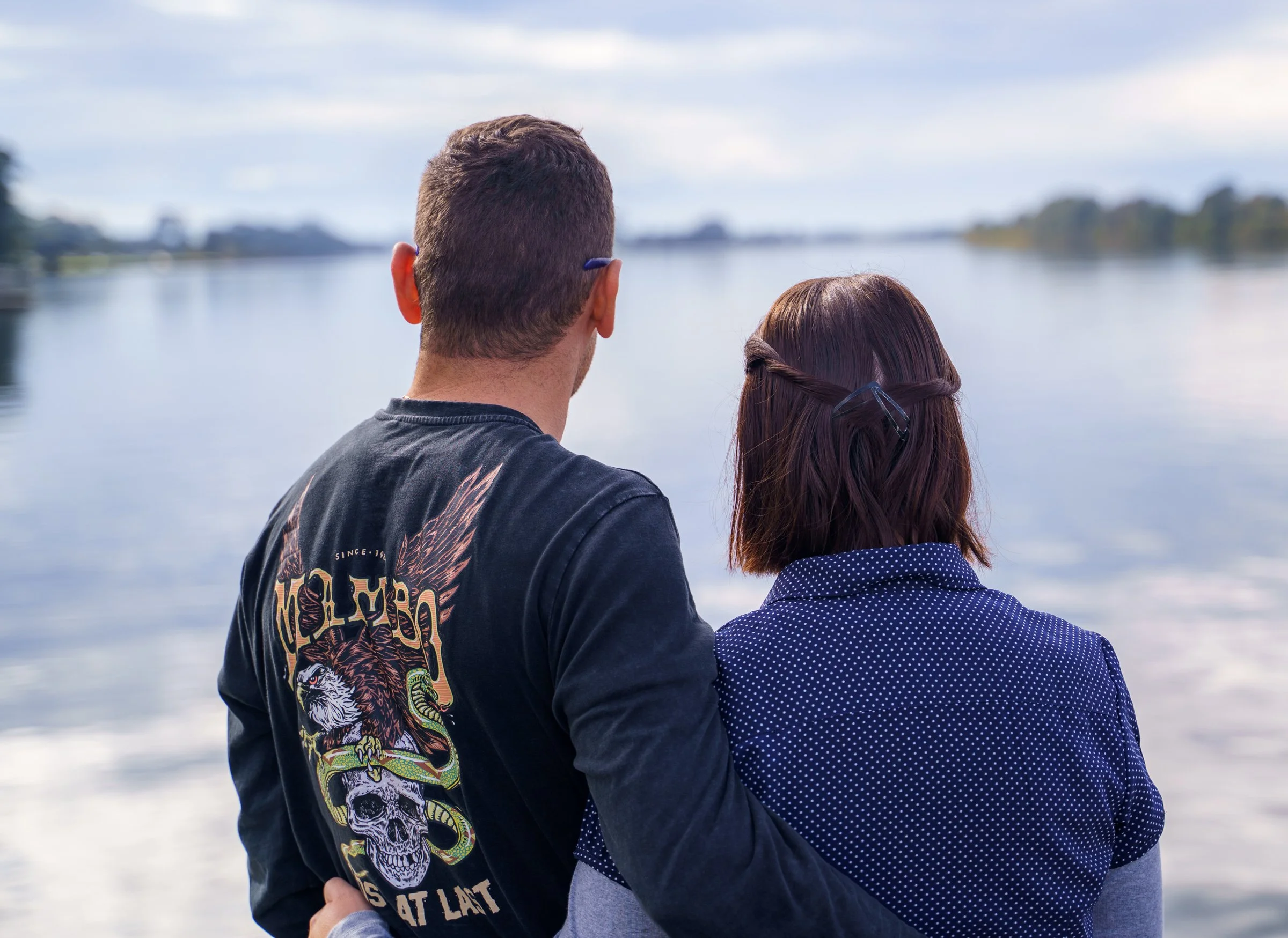 A man and woman standing beside a body of water, with their backs to the camera, overlooking the water and distant shoreline.