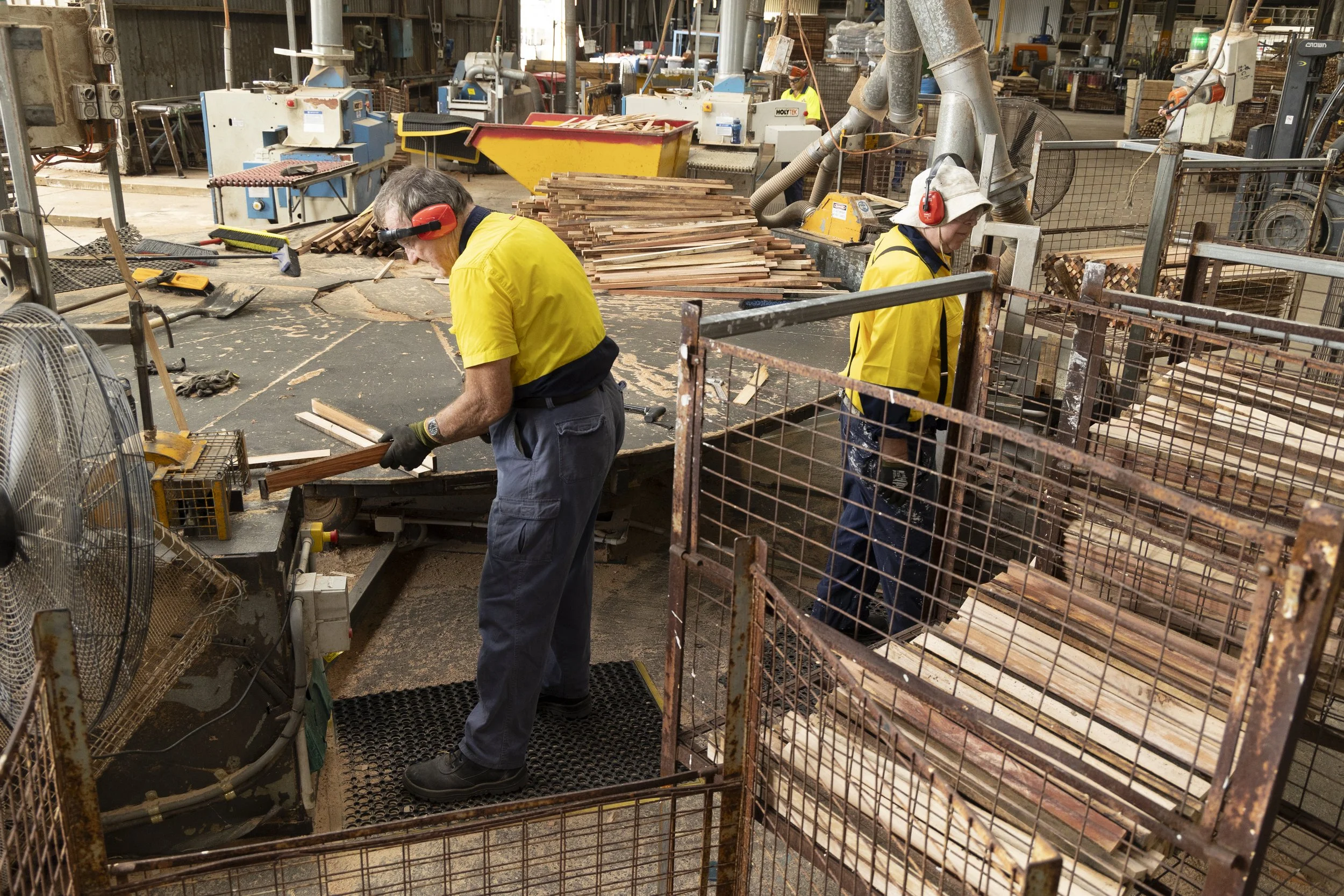 Two workers wearing yellow safety shirts and ear protection are working at Valley Timber. One is handling a piece of wood, and the other is standing near stacked wood planks.