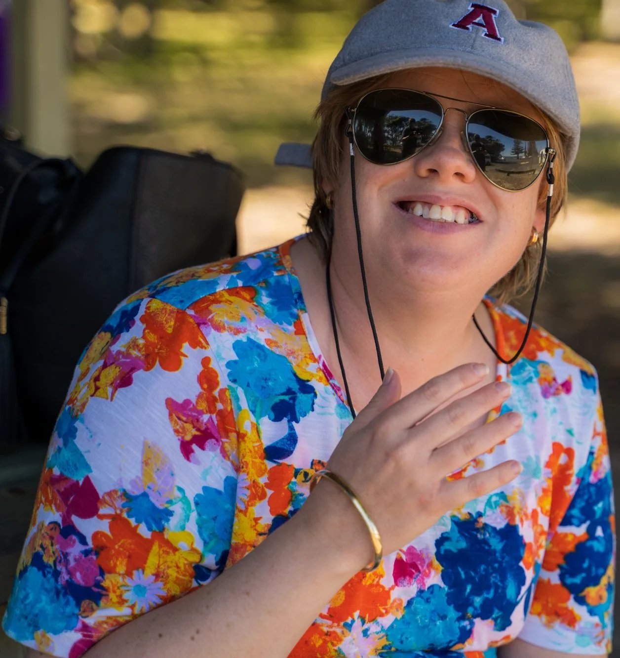 A young woman smiling outdoors wearing a gray baseball cap, sunglasses, and a colorful floral shirt, with a backpack behind her.