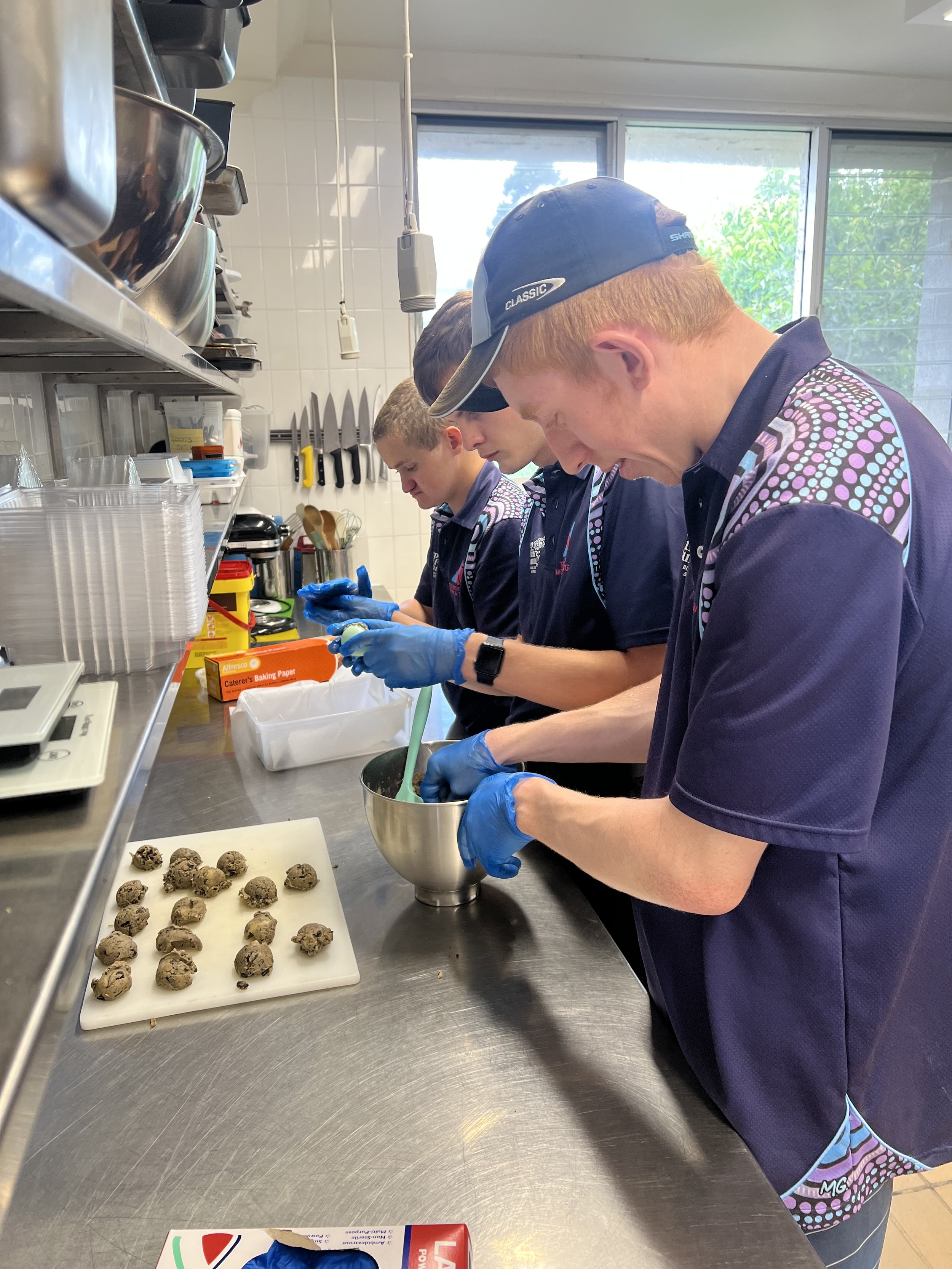 Valley team members working at a stainless steel counter, with a tray of cookie dough balls in front of them. The kitchen has shelves with bowls and knives, and a window letting in natural light.