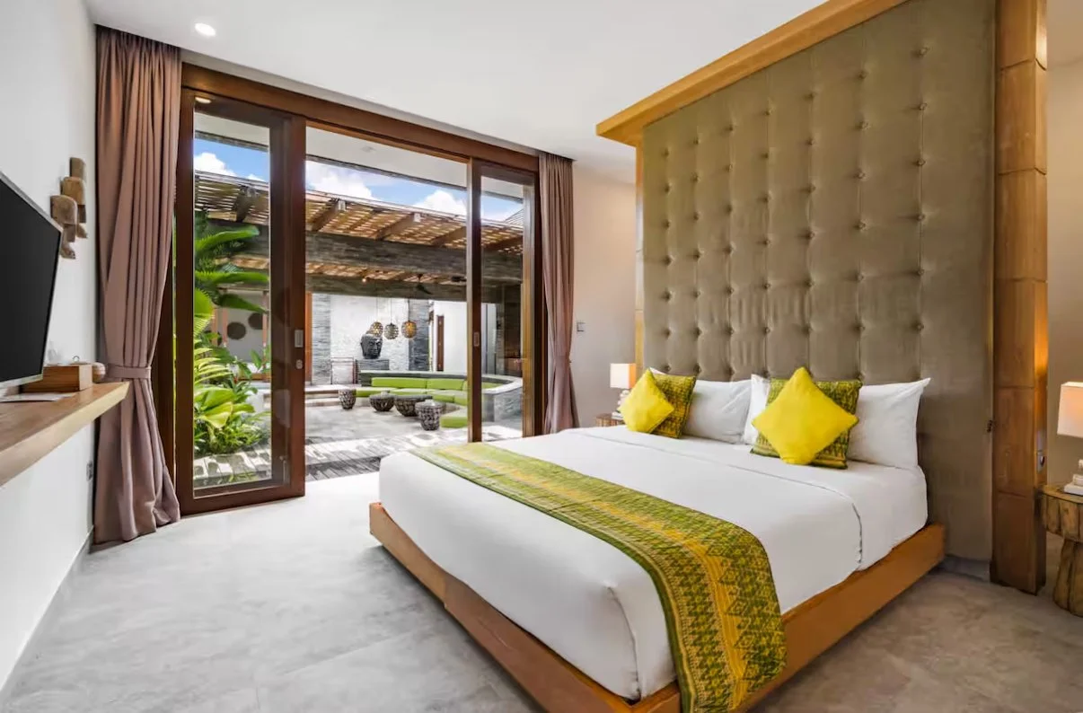 A modern hotel room with a large bed, green and yellow pillows, a wooden headboard, bedside lamps, a flat-screen TV, and a sliding glass door opening to an outdoor patio with seating and lush greenery.
