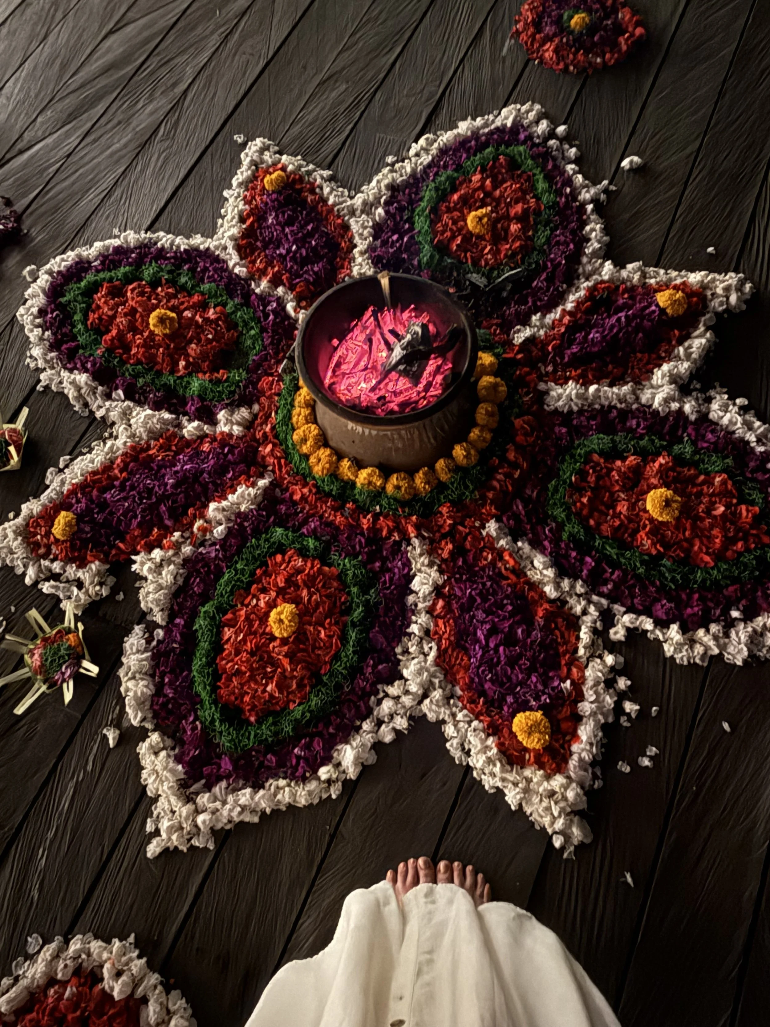 Colorful floral rangoli design with a candle in the center, decorated with marigold flowers, on a dark wooden floor.
