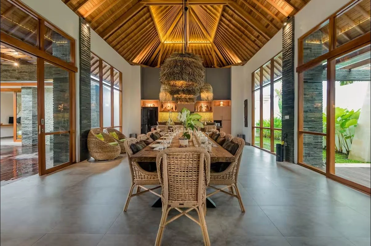 A spacious dining room with a wooden ceiling, large windows, and modern decor, featuring a long wooden dining table with wicker chairs and a central flower arrangement.