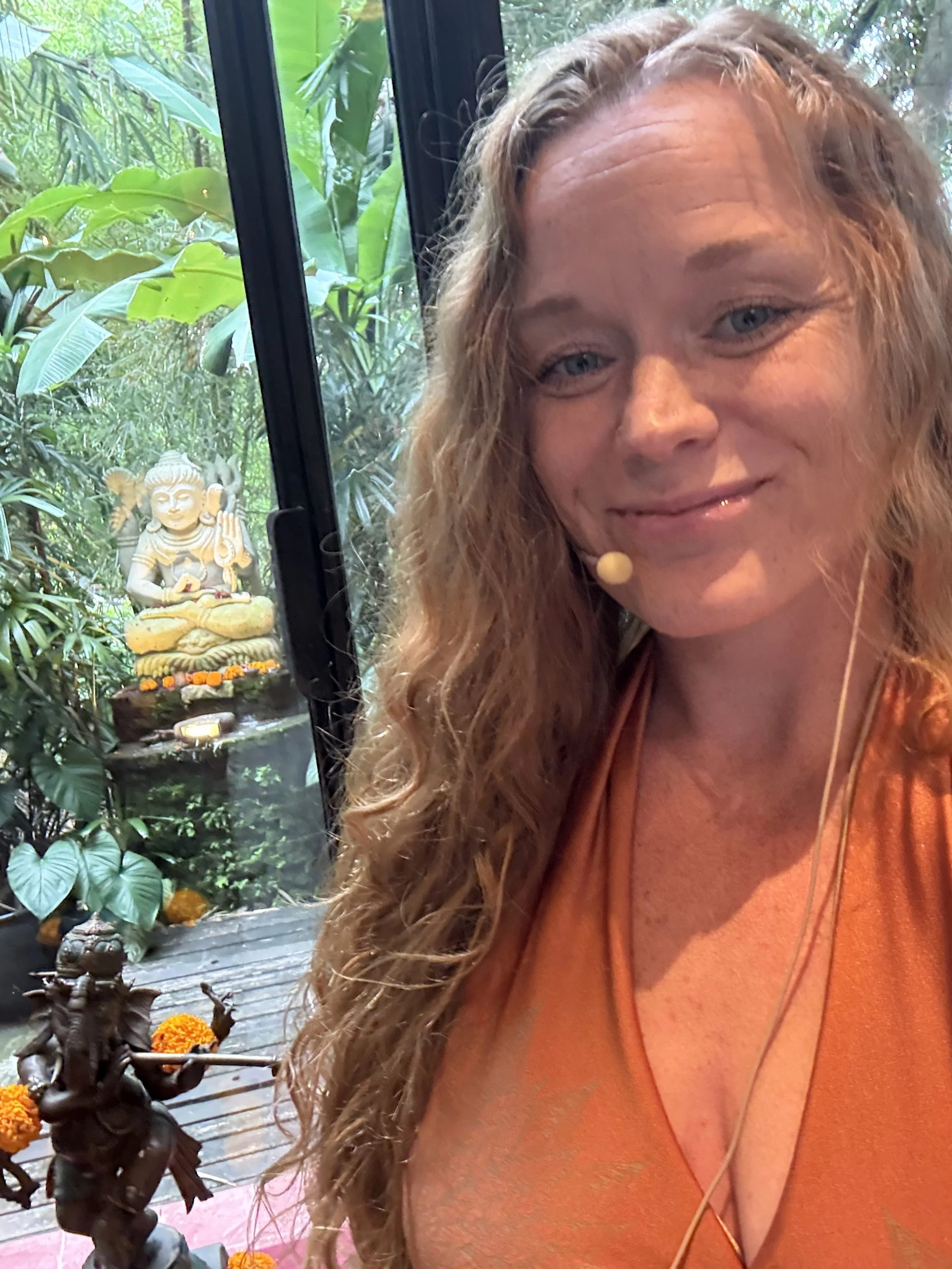 A smiling woman with long curly hair taking a selfie indoors near a lush tropical garden with statues of Hindu deities, including Ganesha, and decorative marigold flowers.