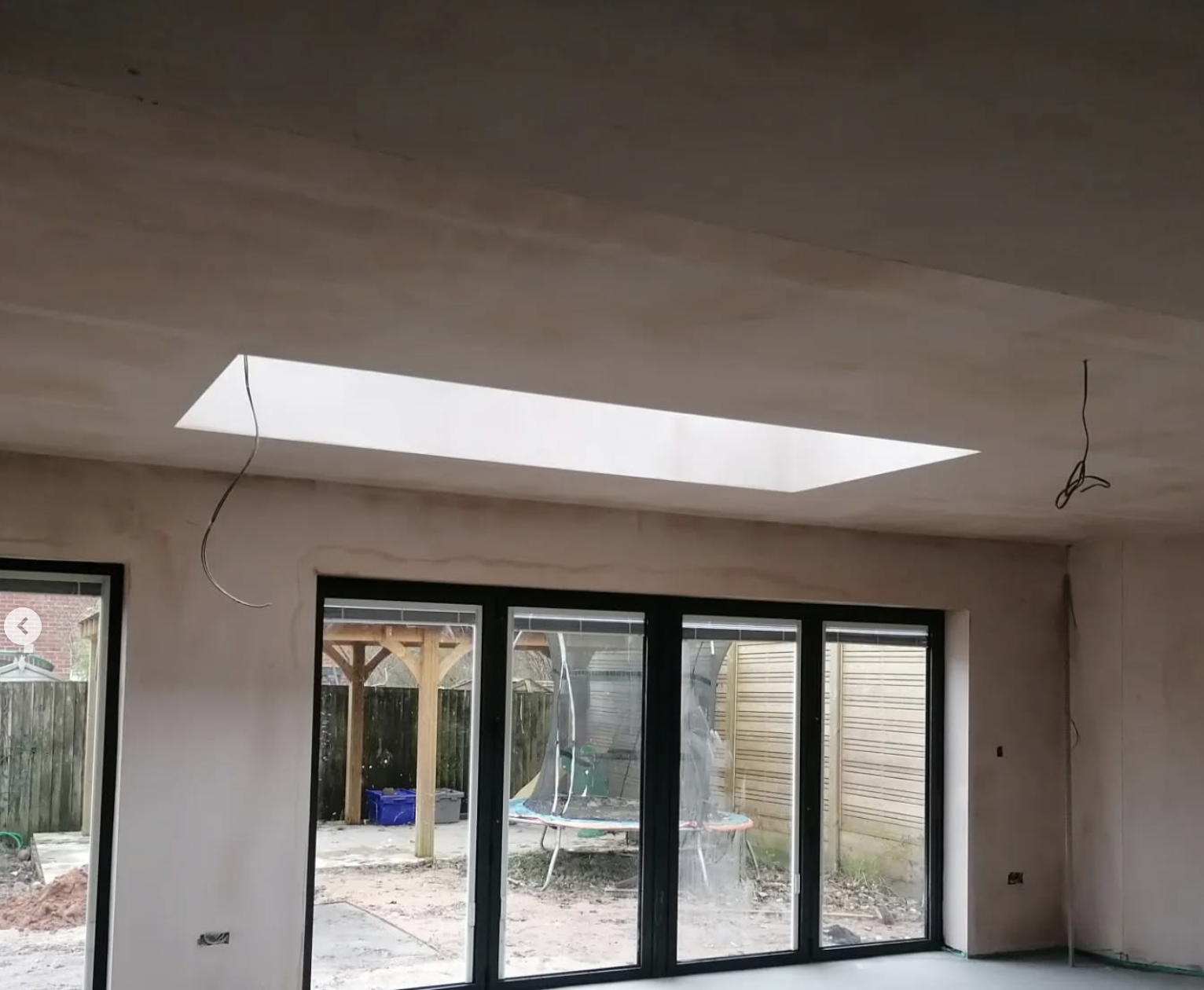 Interior room under construction with a large ceiling skylight and sliding glass doors leading to a backyard with a trampoline and wooden fencing.