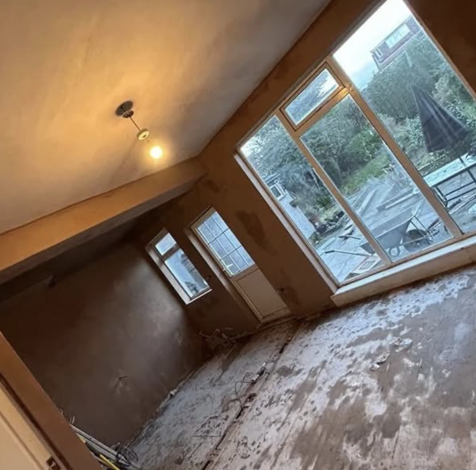 Empty room with large windows, wooden walls, unfinished floor with exposed wires, and a door leading outside, possibly under renovation.