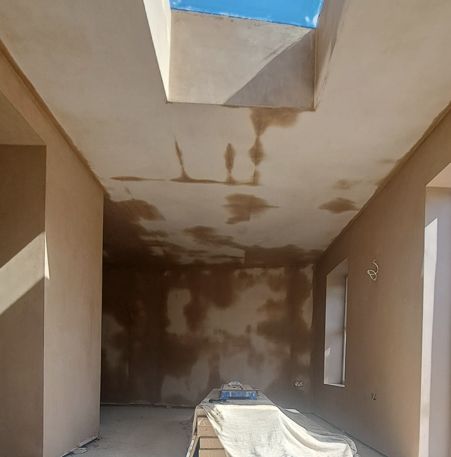 Room under construction with unfinished walls and ceiling, visible window openings, and construction materials on the floor.