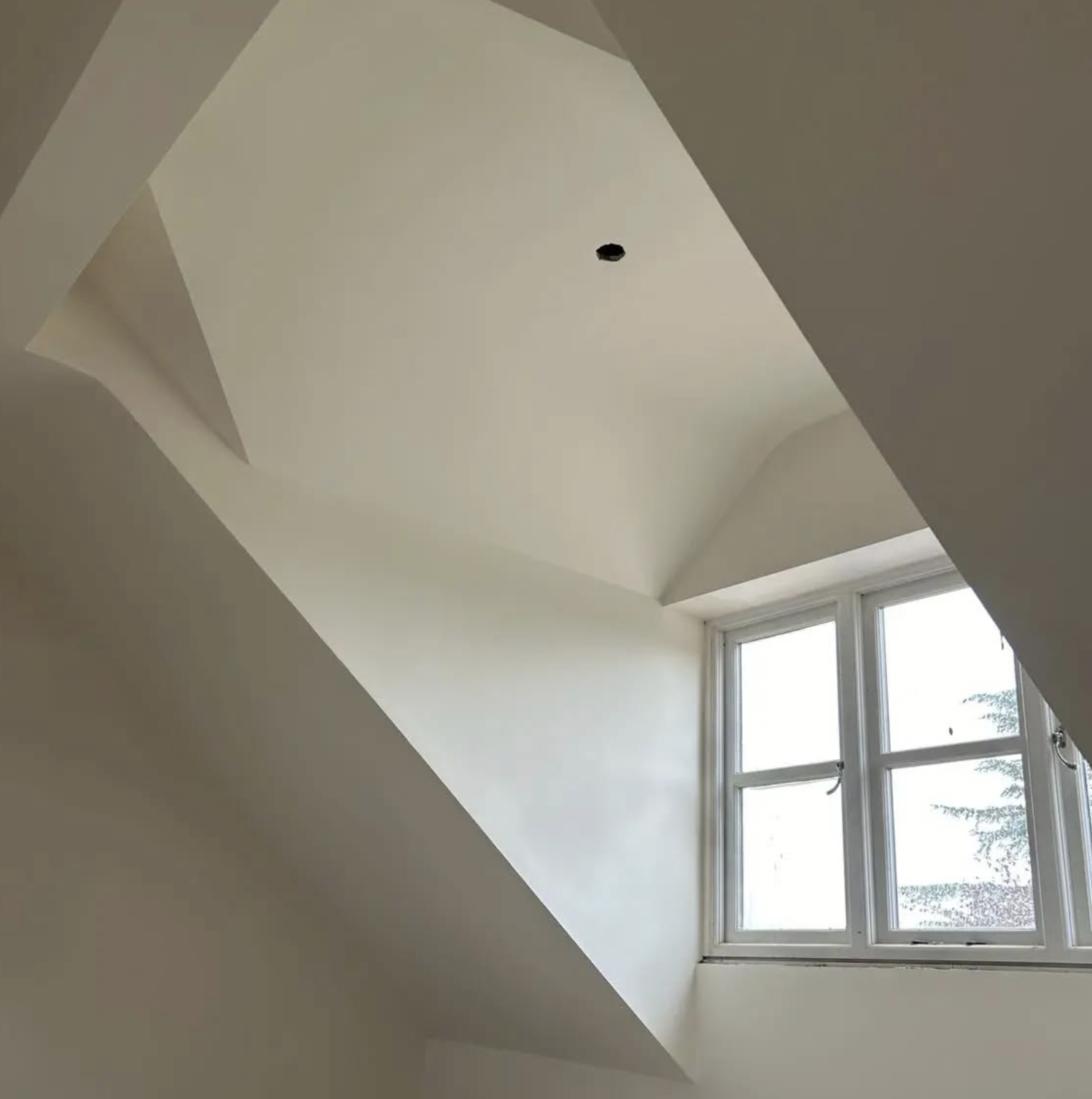 Ceiling with a hole near the center and a window with four panes letting in natural light in a room with neutral-colored walls.