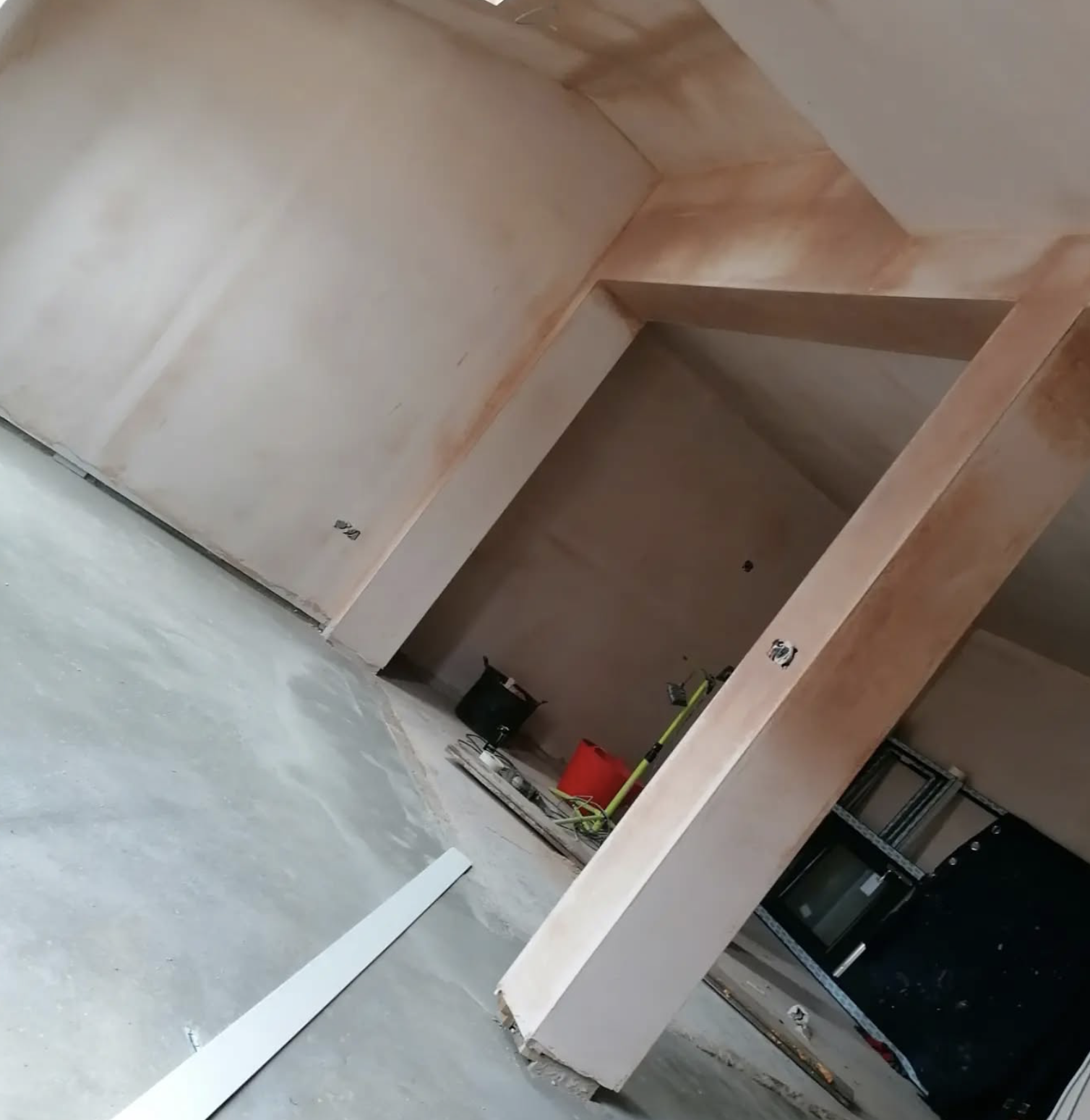 An unfinished room under construction with bare walls and tools on the floor, including a black bucket, a red container, and various construction equipment.