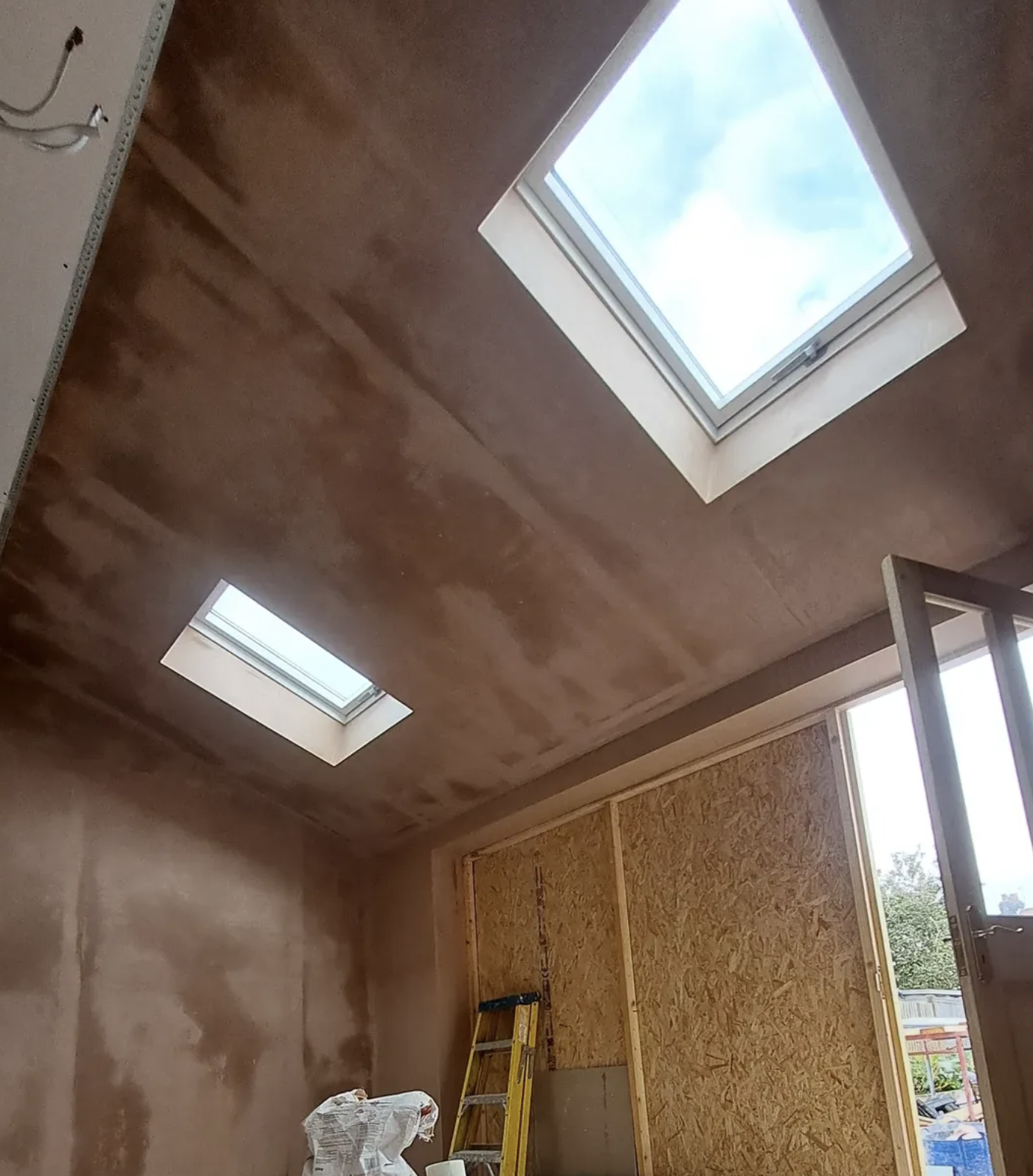 An unfinished room under construction with two skylight windows on a brown-painted ceiling, a ladder, and a partially boarded wall.