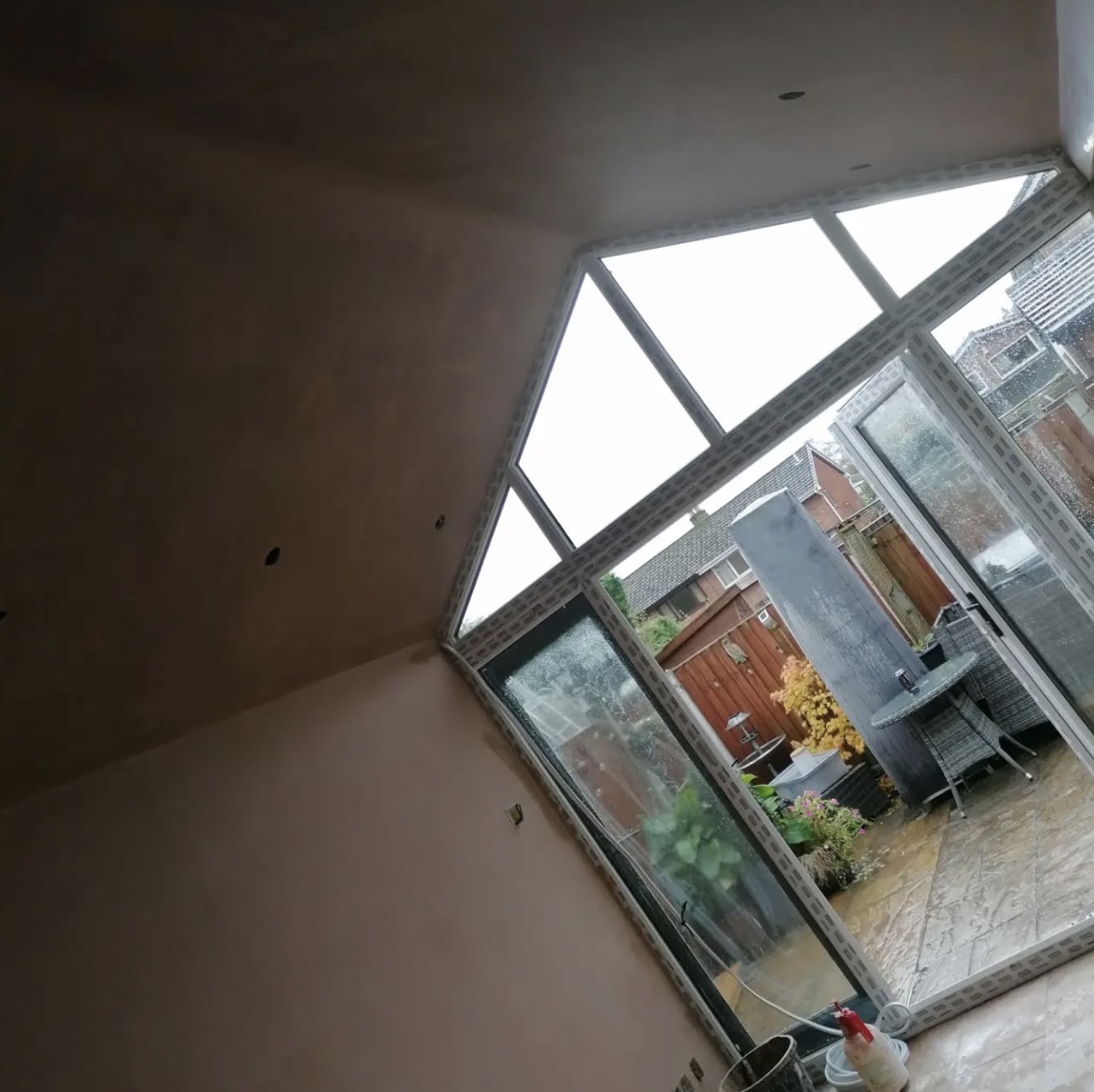 Interior of a room under construction or renovation, with large windows opening to a backyard patio with outdoor furniture and plants, viewed from inside the room with partially painted or unfinished walls.