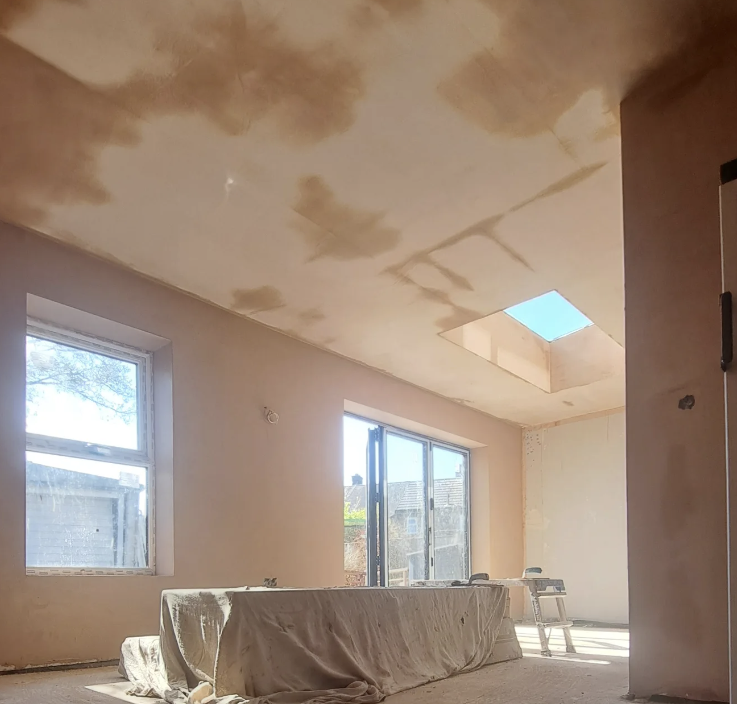 Room under renovation with unfinished walls, a ceiling with visible patches, a skylight, and construction materials including a covered object and a step ladder.