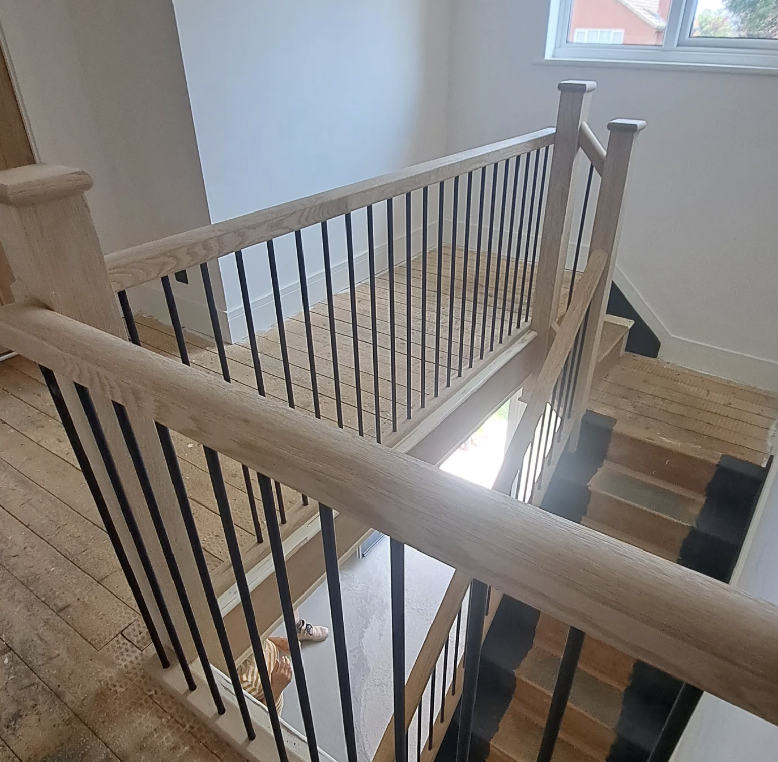 Interior view of a wooden staircase with a balcony railing in a house, featuring wooden steps, black metal spindles, and a window at the top letting in natural light.