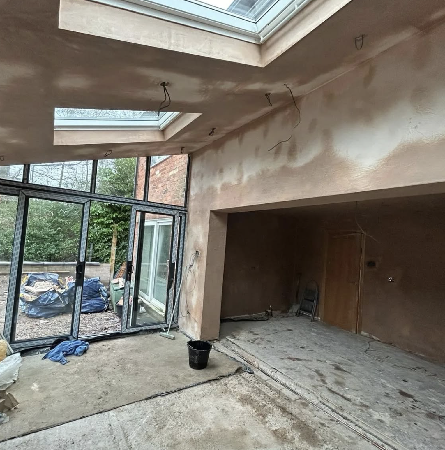 Inside of a house under construction, with unfinished walls, exposed wiring, and a partially installed concrete floor. Large glass sliding doors leading to an outdoor area with construction materials.