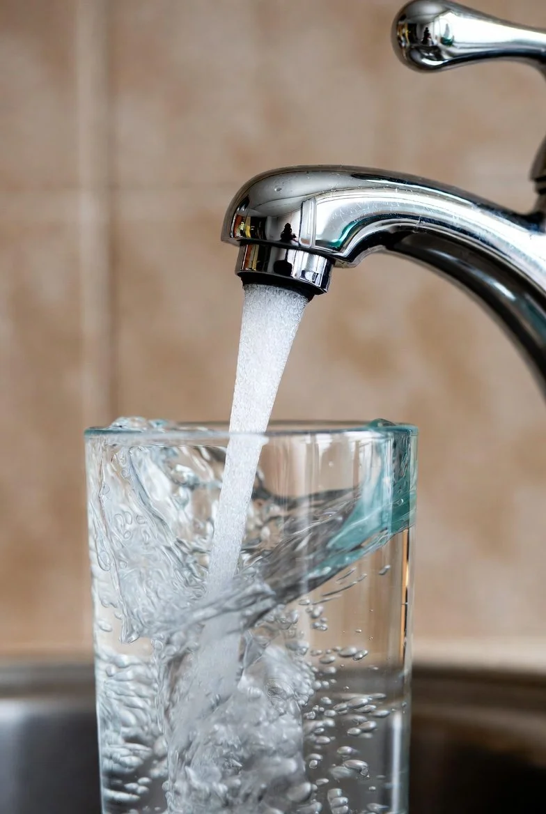 A close-up of a chrome faucet with water running into a clear glass filled with water, against a beige tiled wall background.